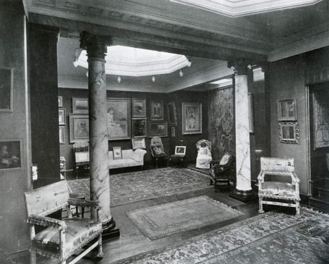 Black and white photograph. Two veined marble columns frame an opening into a space hung with works of art. An octagonal oculus lets in natural light above, and couches and chairs line the walls.