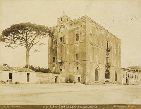 Sepia-toned photograph. We see blocky building with three levels, all with arched doorways or blind arches over windows. A large tree stand directly adjacent.