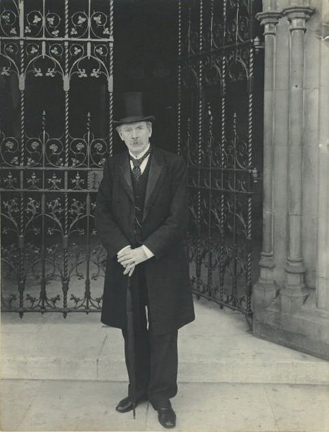 Black and white photograph. Pale-skinned man with a bushy moustache stands in front of an open wrought iron gate. He wears a black suit, coat, and top hat, and he rests his hands on an umbrella held like a cane.
