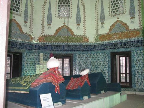 Architecture. A soaring domed space is covered with decoration and patterned tile. A band of tile with Arabic script in white is against cobalt blue runs around the foot of the dome. The tiles on the walls below are densely ornamented with floral designs in blue, red, and white. Four tombs are on a platform in the centre of the space.