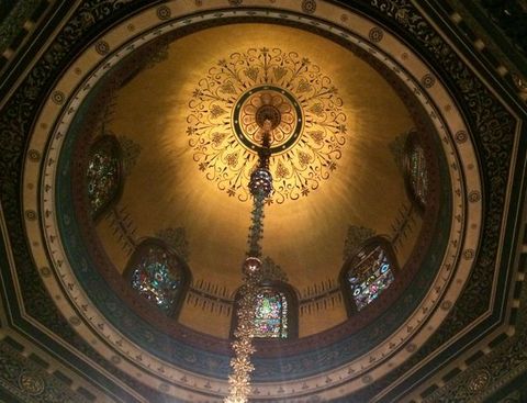 Architecture. We look up into a golden dome lined with arched stained glass windows. The underside of the dome is painted with delicate black patterns to make a medallion around the chain from which a light hangs; the light is out of frame.