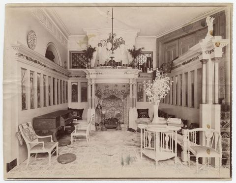Black and white photograph. A hall-like room is lined with tall, narrow panels under an architectural entablature leading to an ornate mantlepiece over a richly decorated fireplace across from us. The floor is also patterned, and delicate white furniture is arranged along the edges of the space.