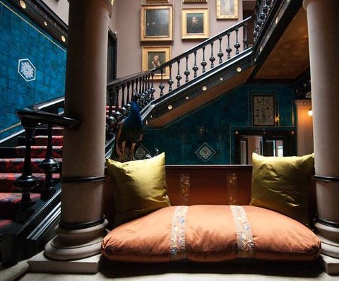 Architecture. Close to us, a cushion and pillows are on a seat between two stone columns. A black wood staircase winds up behind it. Paintings are hung on the wall opposite us, along the top half of the stairs.