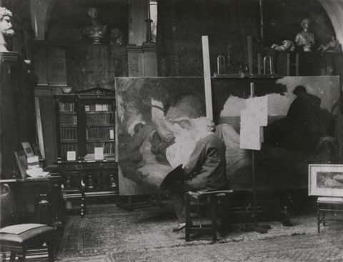 Black and white photograph. A bearded man sits facing away from us as he works on a wide canvas that spans more than half the room. Busts, books, and other material are on shelves behind the painting.