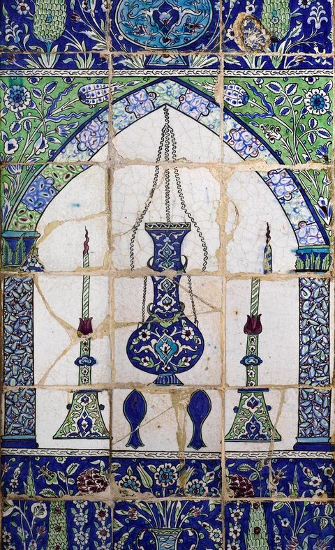 Tiles. Tiled panel shows a round-bellied mosque lamp and two candlesticks under an arch. The objects and patterns on and around the arch are in shades of jewel-toned blue and green.