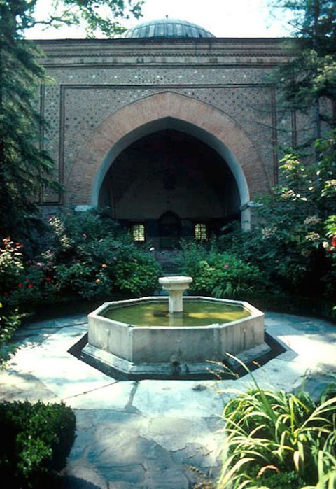 An octagonal fountain is surrounded by a white stone walkway, which is itself encased in plants, shrubs, and trees. An arched opening in the brick wall opposite us leads to a darkened space.