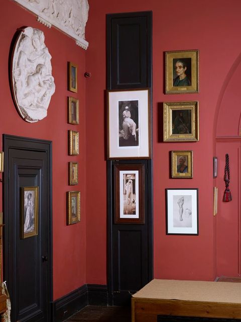 Colour photograph. The corner of a room with muted red walls is hung with paintings, photographs, relief sculptures, and drawings. A black door with a black doorknob is to our left. Just beyond the turning of the corner, a black door is thinner but about twice as tall. Both are closed and works of art hang on both.