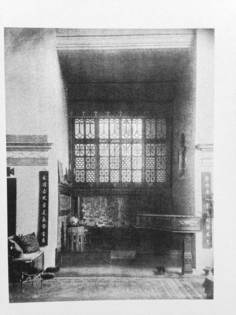 Painting. Two women and a child sit on a couch against a maroon-red wall, which has a line of pictures in gold frames hanging in a row above. The child leans forward and points at something to our right. A large easel or display stand to our right, between us and the women, holds two gold-framed works.