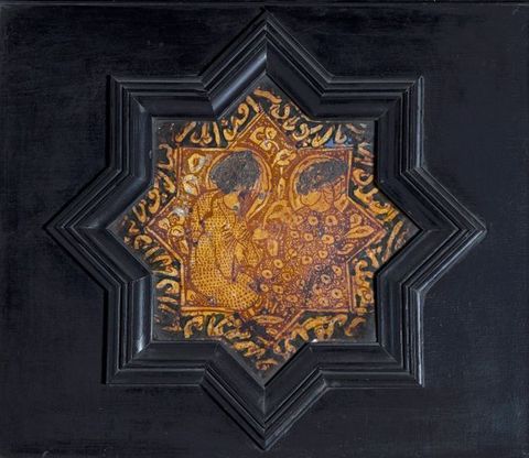 Tile. Eight-pointed tile shows two figures painted in copper red against golden yellow. The black frame follows the contour of the tile and is set into a black wall.
