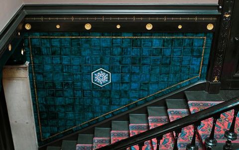 Architecture. From off to the side, we look down a flight of stairs lined with red and blue carpet down the middle of black wood risers. The wall follows the angle down the stairs is filled with vibrant blue tiles around a snowflake-like white design at the centre.