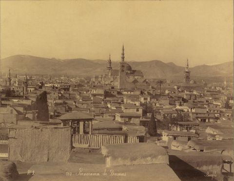 Sepia-toned photograph. A sea of flat rooflines covers the distance to a domed structure with two pointy minarets in the distance. A band of mountains spans the horizon. Text in French along the bottom of the image reads, “No. 2 193. Panorama de Damas Bonfils”.