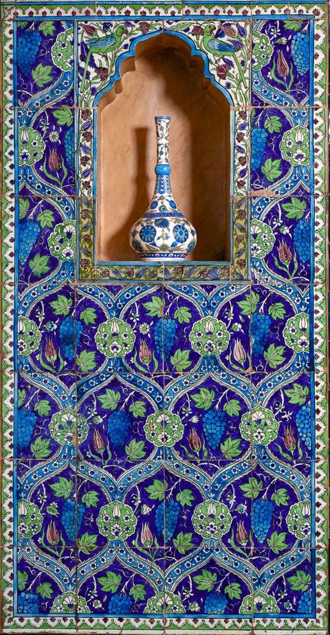 Architecture. Wall tile makes an overarching pattern of continuous S’es surrounding grapes, leaves, and flowers. A blue and white vase with a long, narrow neck and flaring body sits in an arched niche near the top.