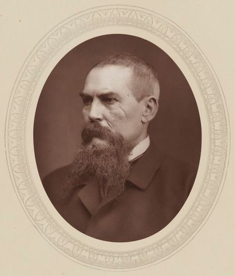 Sepia-toned photograph. In an oval frame, a pale-skinned man sits angled to our left and looking in that direction. His hair is cut short, and he has a long, forked beard. There are wrinkles at the corner of the eye we can see, and a long wrinkle or scar down his cheek.
