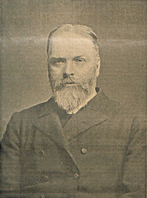 Black and white photograph. Shown from the chest up, a pale-skinned man with a full beard faces us and looks slightly off to our left. He has a low brow over clear eyes and his lips are parted.