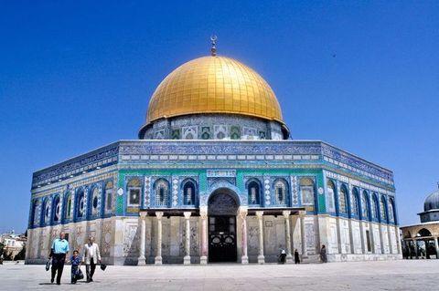 Architecture. A shiny gold dome rises above an octagonal building covered almost entirely in blue tile. The lower half of the octagonal structure is clad in patterns made with white, soft grey, and muted red stone. Eight columns are around the arched entryway, four to each side.