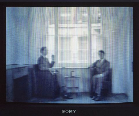 Mixed media. A film still is shown on a black TV set with a “SONY” logo under the screen. Two white men in suits sit in chairs, facing each other in front of a window with curtains pulled back. The still is a grainy texture, in greyscale with very thin vertical striations that lend a fuzzy quality to the image details.